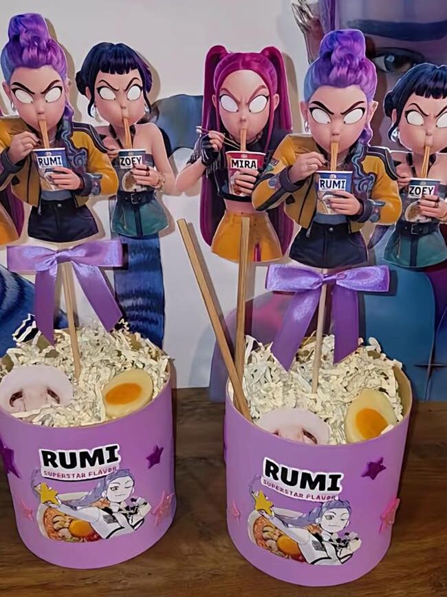 K-Pop Demon Hunter themed party decorations featuring purple ramen bucket props with character cut-outs of Rumi, Zoey, and Mira.