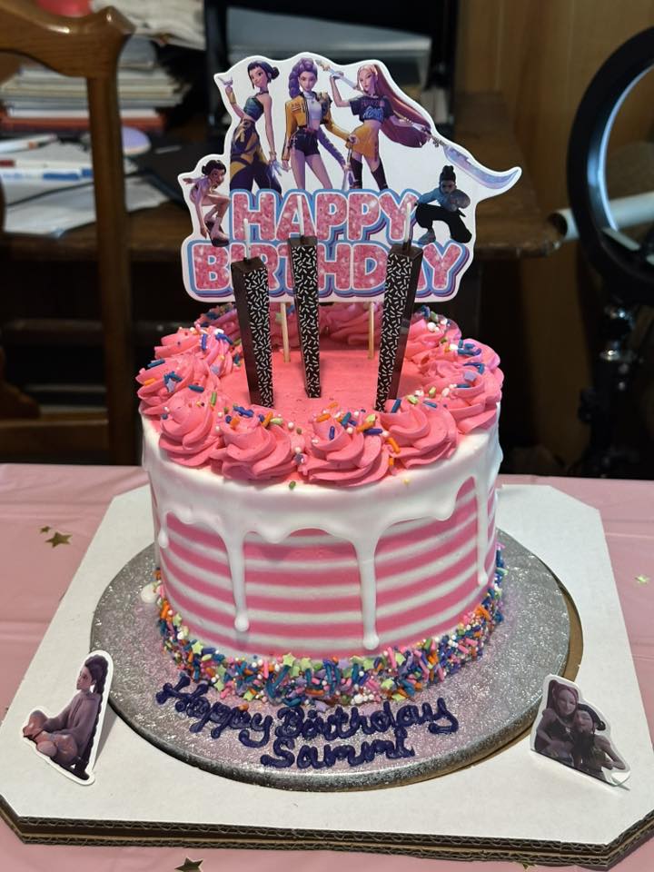 Pink K-Pop Demon Hunter birthday cake with striped icing, character toppers, and sprinkles, perfect for a themed kids’ party.