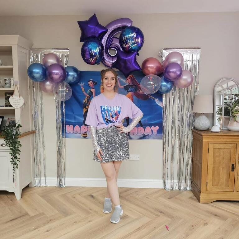 K-Pop Demon Hunter party entertainer in Kent wearing a sparkly outfit and themed top, standing in front of birthday decorations with balloons and a K-Pop Demon Hunter backdrop.